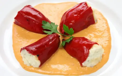 Cuatro pimientos rojos rellenos de bacalao, servidos sobre una cremosa salsa naranja y decorados con perejil.