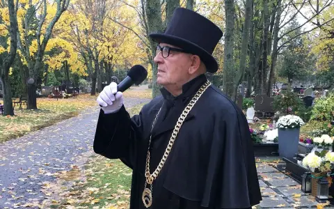 Uroczysty mistrz ceremonii pogrzebowej w czarnym stroju i cylindrze, z mikrofonem w ręku, przemawia na cmentarzu. Cena jego usług jest adekwatna do powagi chwili.