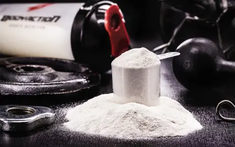 Polvo blanco en una cuchara medidora, junto a pesas y un bote de suplemento. Descubre para qué sirve la creatina y potencia tus entrenamientos.