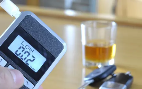 Alkomat z wyświetlaczem pokazującym 0.02% BAC, obok kieliszek z alkoholem i kluczyki samochodowe.