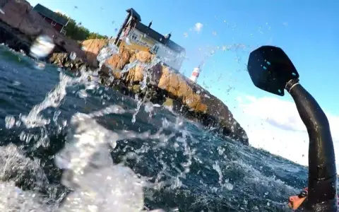 Vattenstänk och en paddel i förgrunden. I bakgrunden en klippa med hus och ett litet fyrhus. En del av en farleden swimrun-deltagare syns.