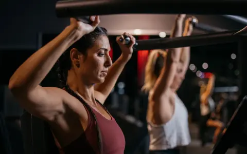 Zwei Frauen trainieren im Fitnessstudio. Sie nutzen eine Maschine, die zeigt, welche Muskelgruppen zusammen trainieren.