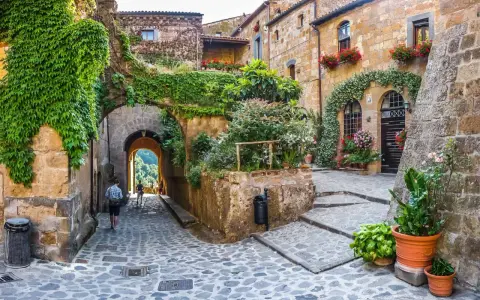 Un vicolo acciottolato con case in pietra ricoperte di edera e fiori. Un uomo cammina verso un arco che si apre su un paesaggio verde. Uno dei tanti posti belli nelle Marche.