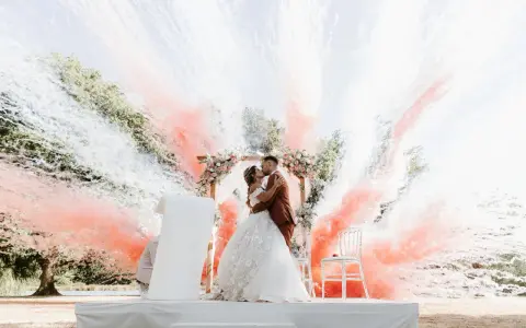 Un couple s'embrasse sous une arche fleurie, entouré de fumée rose et blanche. Ces photos mariage capturent un moment magique.