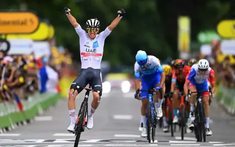 A cyclist in a white UAE Emirates jersey celebrates victory at the Tour de France, arms raised in triumph as he crosses the finish line.