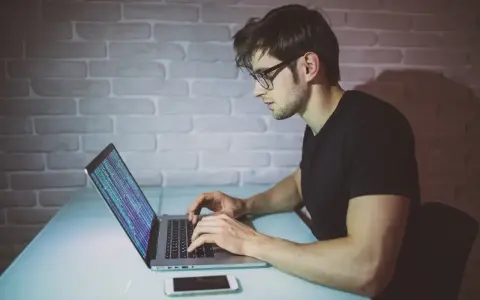 Młody mężczyzna w okularach pisze kod na laptopie. To jego pierwszy krok, by dowiedzieć się, jak zostać informatykiem.