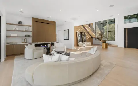 Soggiorno luminoso con camino moderno, divano curvo e poltrone bianche. Scale a vista e grandi finestre definiscono l'arredamento minimal chic.