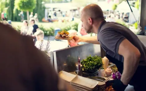 Mann reicht ein Gericht auf einem Holzbrett bei einem Street Food Festival. Die Preise sind hier fair für die leckere Auswahl.