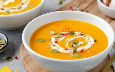 Velouté de courge butternut orange servi dans un bol blanc, garni de crème, de graines de courge et de poivre rose.