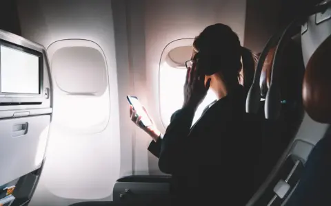Kobieta w okularach trzyma telefon, zastanawiając się, czy w samolocie jest wifi. Obok ekran rozrywki.