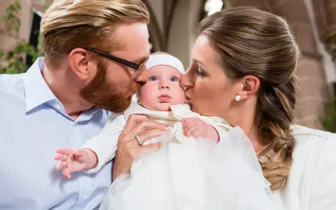Eltern küssen ihr Baby bei der Taufe. Die Frage, wie viele Paten darf man haben, beschäftigt viele.