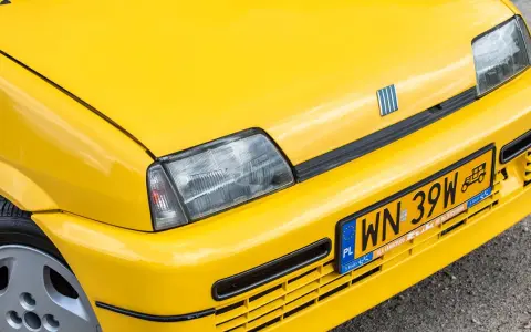 Żółte tablice rejestracyjne na błyszczącym, żółtym samochodzie Fiat. Widoczny fragment maski, reflektor i zderzak.