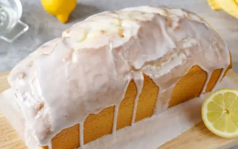 Ein saftiger Zitronenkuchen in Kastenform, überzogen mit süßem Zuckerguss. Ein Zitronenviertel liegt daneben.
