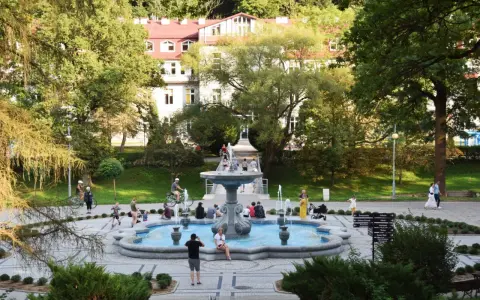 Rymanów Zdrój atrakcje: ludzie odpoczywają przy fontannie w parku zdrojowym, w tle budynek uzdrowiskowy.