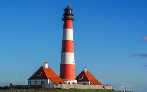 Wysoka, czerwono-biała latarnia morska w Jastarni strzeże wybrzeża. Obok stoją dwa domki z pomarańczowymi dachami.
