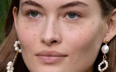 Primer plano de un rostro femenino con pecas naturales salpicando su piel. Lleva pendientes de perlas colgantes.