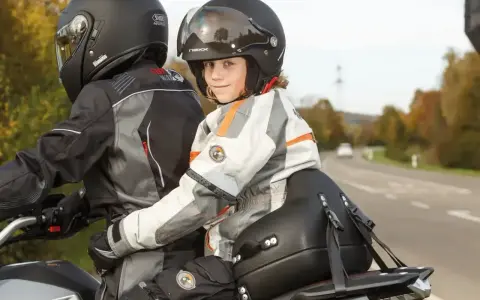 Uśmiechnięte dziecko na motocyklu, w kasku i kurtce, siedzi za kierowcą.