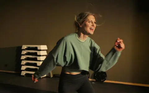 Glad kvinna med blont hår som springer intervaller i ett gym. Hon har på sig en grön tröja och svarta träningsbyxor.