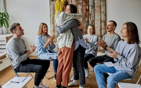 Grupa ludzi siedzi w kręgu, klaszcząc. Dwie osoby w centrum mocno się przytulają. Co to aa? Wsparcie i akceptacja.