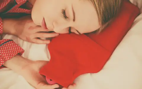 Une femme dort paisiblement, blottie contre une bouillotte rouge. Une astuce pour savoir comment faire une bouillotte avec une serviette pour se réchauffer.