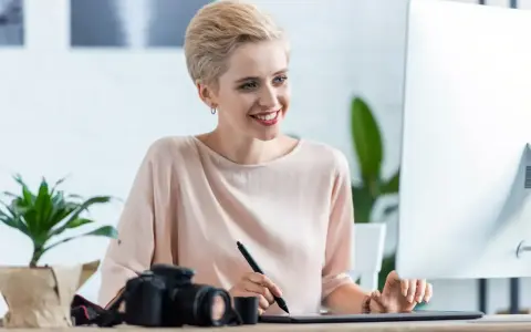 Uśmiechnięta kobieta z krótkimi blond włosami pracuje przy komputerze, obok aparat fotograficzny. Sprawdza najnowsze oferty pracy w Pile.