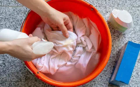 Ręce rozpylają płyn na poplamioną tkaninę w pomarańczowym wiadrze z wodą.