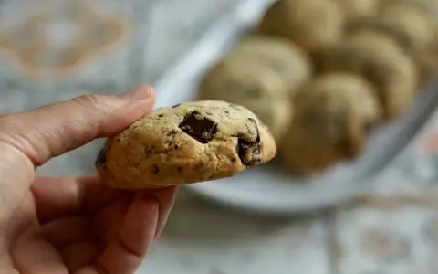 Une main tient un cookie épais et moelleux aux pépites de chocolat. D'autres cookies sont disposés sur une assiette en arrière-plan.