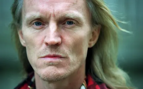 Närbild på John Holm med blå ögon och blont hår. Hans ansikte bär spår av livserfarenhet, en sordinerad blick.