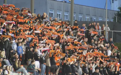 Tłum kibiców na stadionie z szalikami w barwach pomarańczowo-czarnych. Widać napisy "Chrobry" i "Chrobry Głogów".