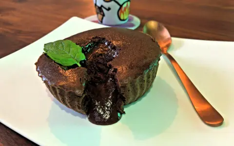 Un délicieux mi cuit chocolat coulant, garni d'une feuille de menthe, sur une assiette blanche. Une cuillère dorée est à côté.