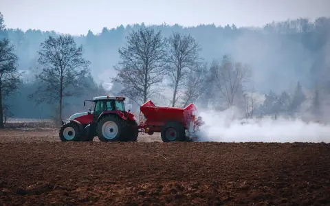 Czerwony traktor z przyczepą rozsypuje nawóz na polu. To przygotowanie gleby, by warzywa lubiące wapnowanie, jak pomidory czy kapusta, dobrze rosły.