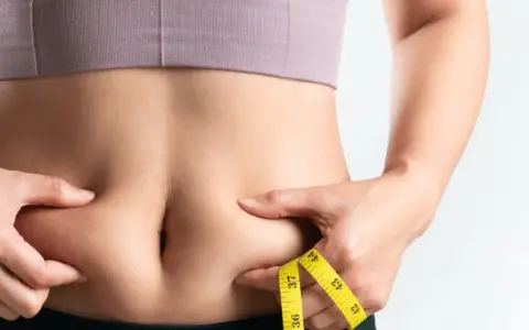 Frau kneift Bauchfett, Maßband zeigt Umfang. Tipps für 10 Hausmittel gegen Bauchfett.