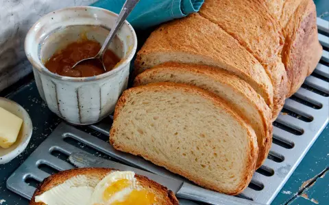 Ein Laib **sauerteig Toastbrot** ist aufgeschnitten. Daneben stehen Marmelade, Butter und ein Messer mit einem Stück Brot, das bestrichen ist.