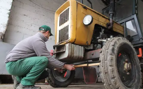 Mężczyzna w zielonych spodniach i czapce reguluje przednie koło żółtego ciągnika 4x4, pokazując, jak ustawić zbieżność.