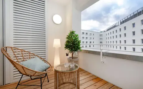 Przytulny balkon z drewna, z wiklinowym fotelem, stolikiem i rośliną. W tle białe budynki.