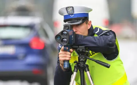 Policjant w kamizelce odblaskowej mierzy prędkość samochodu za pomocą radaru. Trwa dozór policyjny na drodze.