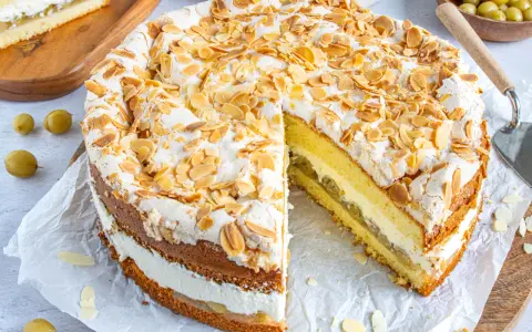 Ein köstlicher Stachelbeer Baiser Kuchen mit Biskuitböden, Sahne und Mandelsplittern. Ein Stück ist bereits herausgeschnitten.