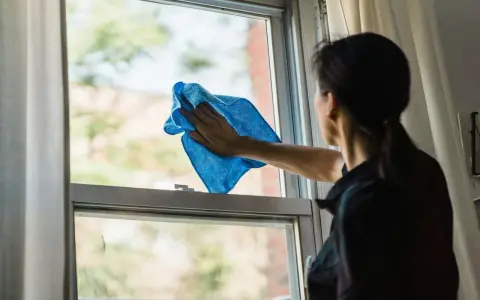 Kobieta myje okno niebieską ściereczką. Dowiedz się, czym myć okna, by lśniły czystością.