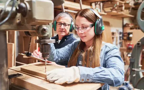 Junge Frau lernt an einer Bohrmaschine, während ein älterer Mann zusieht. Eine gute Ausbildung, welche Ausbildungen dauern 2 Jahre, ist hier vielleicht der Anfang.