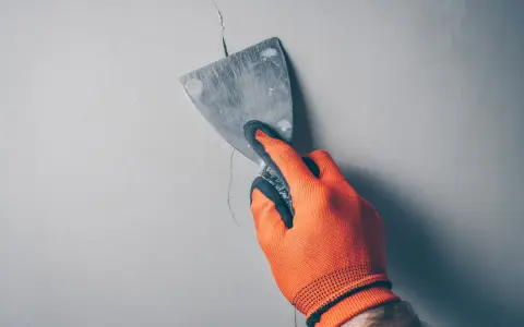 Ręka w pomarańczowej rękawicy zacierająca pęknięcie w ścianie szpachlą. Idealne rozwiązanie, czym wypełnić głębokie pęknięcia w ścianie.