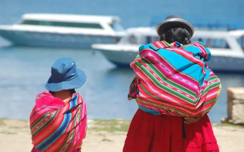 Zwei cholitas escaladoras, gekleidet in bunte Ponchos und Hüte, blicken auf Boote auf einem See.