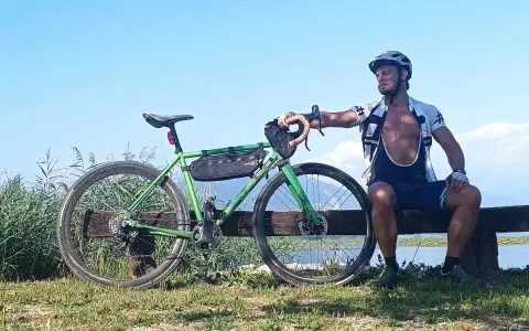 Ciclista riposa accanto alla sua bici verde, pensando alle tabelle per dimagrire in bicicletta. Cielo azzurro e lago sullo sfondo.