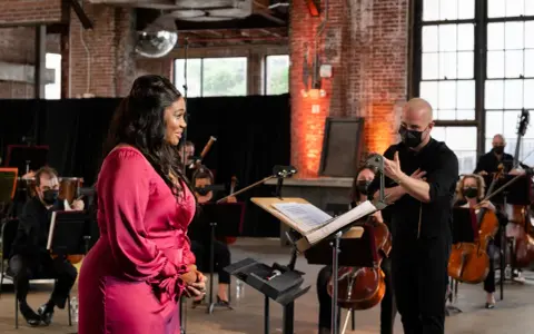 Une chanteuse en robe rose discute avec un chef d'orchestre masqué, entourée de musiciens. Elle cherche peut-être à trouver un agent pour sa carrière.