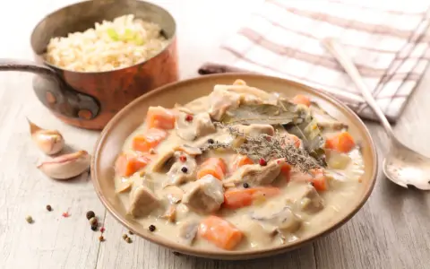 Un plat de blanquette de veau crémeuse avec des carottes, des champignons et des herbes, servi avec du riz dans une casserole en cuivre.