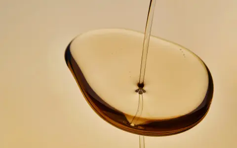 Un filet d'huile de karanja, d'une couleur ambrée, tombe sur une surface liquide, créant des reflets sombres. Attention, l'huile de karanja peut présenter un danger.