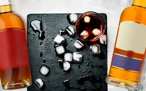 Dwie butelki, jedna z czerwoną etykietą, druga z beżową i fioletową, stoją obok szklanki z lodem i ciemnym płynem. Koniak czy whisky?