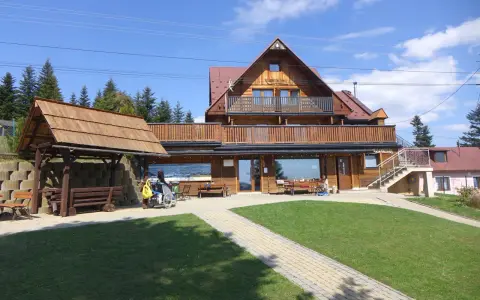 Drewniana bacówka na obidzy z balkonem i tarasem, otoczona zielenią i ścieżkami. Obok wózek inwalidzki z żółtą torbą.