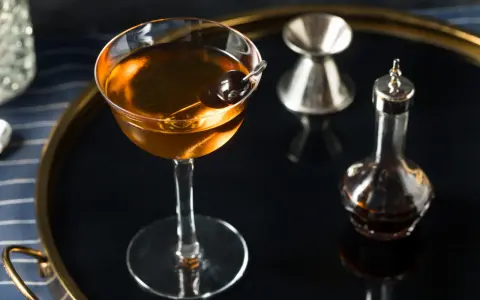Elegancki koktajl w kolorze calvados, z wiśnią, obok kieliszka do mierzenia i buteleczki z bitters. Idealny do deserów.