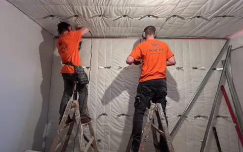 Dwóch mężczyzn w pomarańczowych koszulkach montuje materiał izolacyjny, pokazując, jak wygłuszyć ścianę.