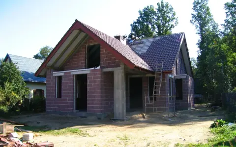 Dom w stanie surowym otwartym, z czerwonej cegły, z częściowo położonym dachem. Wokół plac budowy.